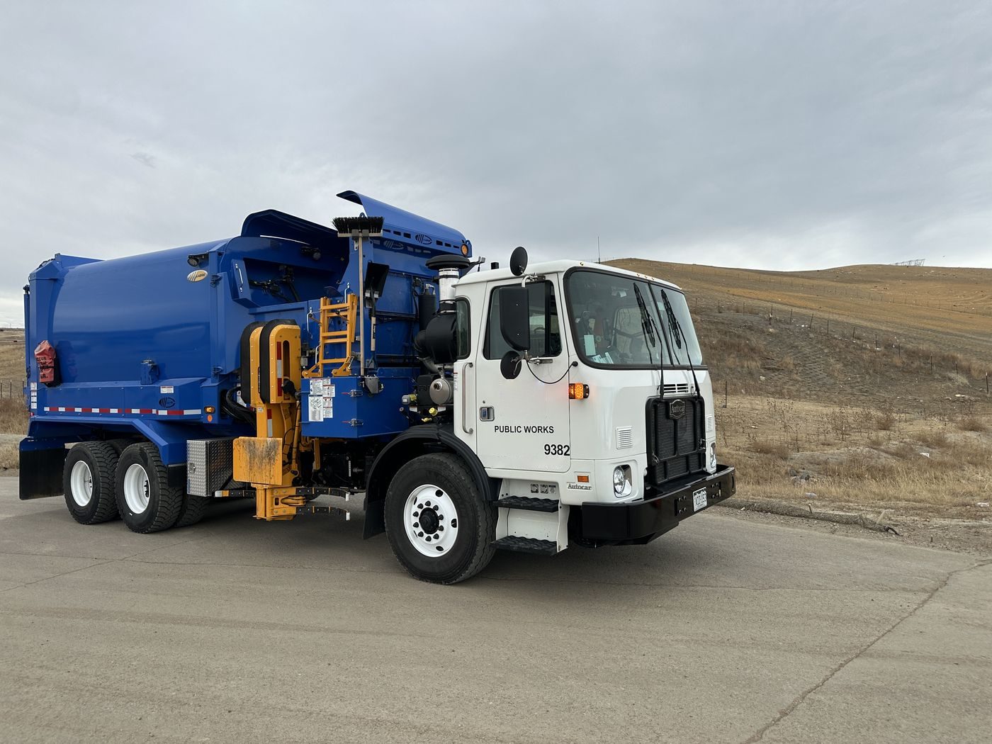 A city public works automated side-load refuse truck parked on a road at the edge of town